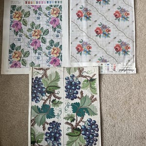 Pode incluir: Três folhas de padrões de ponto cruz com desenhos florais e de frutas. Duas folhas exibem buquês de rosas em rosa, amarelo e vermelho sobre uma grade. Uma folha mostra dois painéis verticais de cachos de uvas roxas com folhas verdes.