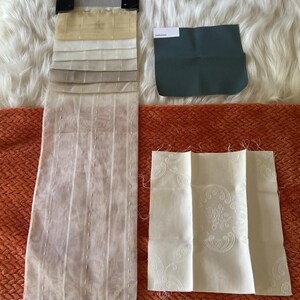 May include: A vertical fabric sample strip featuring various shades of sheer beige and cream with vertical stripes, a single dark teal fabric swatch with a white label, and a square cream fabric swatch with a subtle white floral damask pattern.