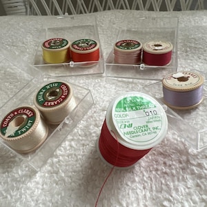 Pode incluir: Uma coleção de carretéis de linha de costura incluindo vários carretéis Coats & Clark's Silk Twist em amarelo, laranja, rosa, vermelho e creme, juntamente com um carretel grande de linha de seda Clover 100% em vermelho e um carretel lavanda, todos em estojos transparentes.