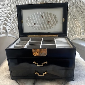 May include: A black jewelry box with a glossy finish, featuring two drawers with gold-toned geometric handles. The interior is lined with light gray fabric, including ring slots and compartments. The lid has a mirror with an oval etched design.