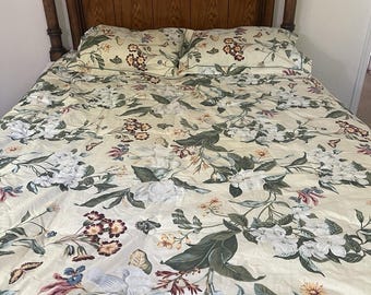 Puede incluir: Una cama con cabecero de madera de parte superior curva y paneles verticales. La cama está cubierta con una sábana de color crema y dos fundas de almohada a juego, decoradas con un estampado botánico de flores blancas, hojas verdes y mariposas.