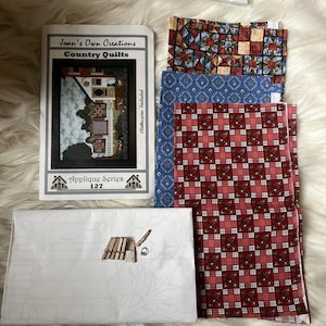 Op de afbeelding: Een knutselpakket getiteld Joan's Own Creations Country Quilts Applique Series 127, inclusief een gedrukt instructieboekje, drie rechthoekige stoffen lappen met geometrische en bloemmotieven, een wit patroonblad en kleine houten wasknijpers.