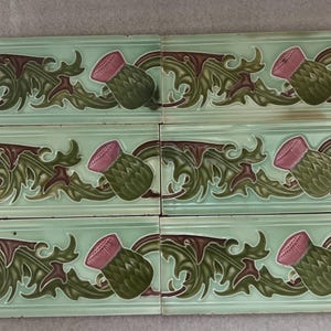 May include: A set of six rectangular ceramic tiles featuring a raised relief pattern of green thistle leaves and pink thistle flower heads against a light mint green background, arranged in two columns of three tiles each with a glossy finish.
