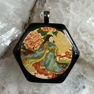 Può includere: Un ciondolo esagonale con una cornice nera lucida e un'illustrazione circolare centrale di una figura in tunica blu che tiene un ventaglio, circondata da peonie rosa e accenti dorati, con un gancio in metallo sulla parte superiore.