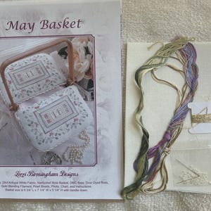 May include: A cross stitch kit titled May Basket by Lorri Birmingham Designs. The kit includes a pattern sheet with a photo of a finished basket, 28ct antique white fabric, assorted green and purple embroidery floss, gold blending filament on a spool, and a small bag of white pearl beads and needles.