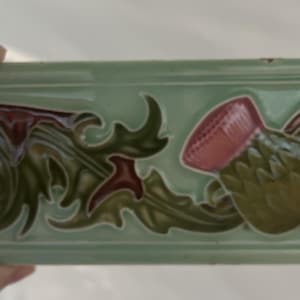 May include: A rectangular ceramic tile featuring a raised relief design of a pink thistle flower and dark green acanthus leaves against a light sage green background. The tile has a smooth, glossy finish and a decorative border along the top and bottom.