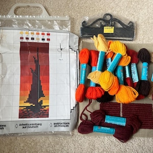 Pode incluir: Um kit de ponto de agulha com um saco plástico contendo o padrão impresso de um veleiro ao pôr do sol, rotulado LITEN SEILBÅT 6605. O kit inclui uma tela bege, um cabide de metal preto e vários novelos de lã Anchor em tons de vermelho, laranja, amarelo e castanho.
