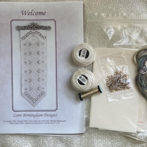 Op de afbeelding: Een kruissteekpakket van Lorri Birmingham Designs genaamd Welcome, inclusief een gedrukt patroonblad, twee witte garenklosjes, een gouden metallic garenklosje, een zakje kralen, een stuk crèmekleurige stof en een bundel gemêleerd borduurgaren.
