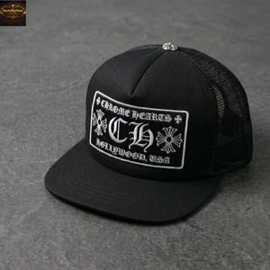 May include: Black trucker hat with a mesh back and a curved brim. The front panel features a white embroidered patch with the text "Chrome Hearts Hollywood, USA" and decorative floral elements. The hat has a silver button on top.