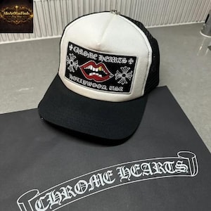 Puede incluir: Una gorra de camionero en blanco y negro con un parche rectangular que incluye el texto Chrome Hearts, Hollywood, USA y un gráfico de labios rojos con dientes. La gorra tiene una visera negra, un panel frontal blanco y malla negra.