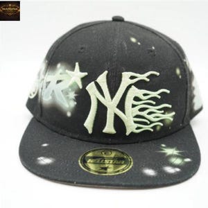 May include: Black baseball cap featuring the New York Yankees logo in a light green color. The cap has a flat brim and a fitted design. The cap has a star and spray paint design on the side and brim.