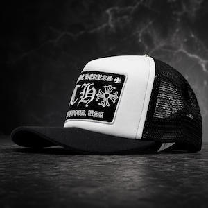 Puede incluir: Una gorra de camionero en blanco y negro con un parche rectangular negro en el panel frontal. El parche muestra el texto blanco CHROME HEARTS, un símbolo de cruz y el texto HOLLYWOOD, USA en fuente gótica. La gorra tiene visera negra y malla.