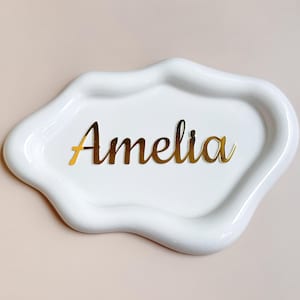 Könnte beinhalten: Eine weiße, unregelmäßig geformte, glänzende Keramikschale mit erhöhten, wellenförmigen Rändern. In der Mitte der Schale befindet sich der Name Amelia, der in einer reflektierenden, metallisch goldenen Schreibschrift auf der glatten Oberfläche steht.