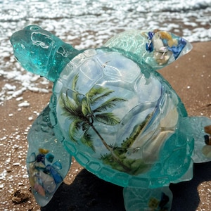 Puede incluir: Una figura de tortuga de resina turquesa con una escena de playa tropical en el interior del caparazón. La tortuga está en una playa arenosa con el océano al fondo. Las aletas de la tortuga están adornadas con piedras de colores.