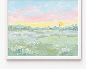 May include: A framed landscape painting in a white frame depicting a golf course at sunset. The scene features a green putting area with a yellow flag, rolling hills in the distance, and a sky filled with soft pastel pink, blue, and yellow hues.