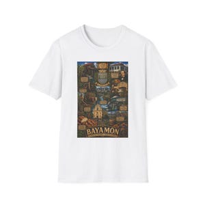 May include: A white short-sleeve t-shirt featuring a detailed, multi-colored graphic print on the chest. The design displays a collage of historical landmarks, a coat of arms, and the text BAYAMON, along with smaller descriptive captions.