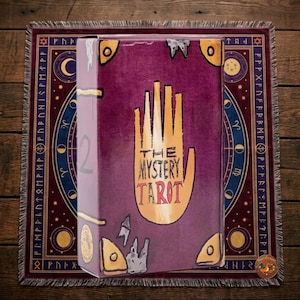 Könnte beinhalten: Eine rechteckige Schachtel für das The Mystery Tarot Deck mit einem violetten Einband, einer großen gelben Handillustration, goldenen Ecken und dem Text The Mystery Tarot. Die Schachtel liegt auf einem quadratischen Tuch mit Himmelsmotiven.