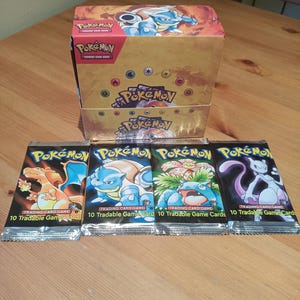 Peut inclure: Une boîte de boosters Pokémon Trading Card Game avec Blastoise sur le couvercle, accompagnée de quatre paquets scellés. Chaque paquet présente un personnage différent : Charizard, Blastoise, Venusaur et Mewtwo, avec 10 Tradable Game Cards.