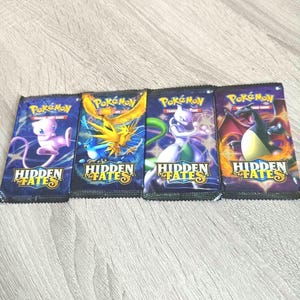 Può includere: Quattro bustine sigillate del gioco di carte collezionabili Pokémon Hidden Fates disposte in fila. Ogni bustina presenta un'illustrazione unica di un personaggio, tra cui Mew, Zapdos, Mewtwo e Charizard, con il logo Hidden Fates stampato in basso.