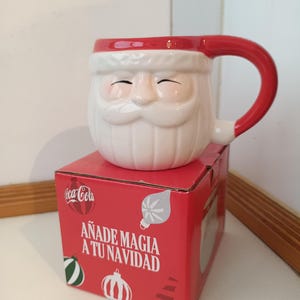Puede incluir: Una taza de cerámica blanca con forma de Papá Noel, con asa y borde rojos, colocada sobre una caja roja. La caja presenta el logotipo de Coca-Cola y el texto en español: "Añade Magia a Tu Navidad" (Añade Magia a tu Navidad).