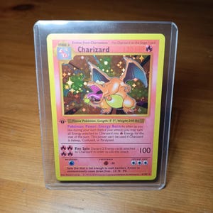 May include: A Charizard Pokémon card in a protective sleeve. The card features a holographic image of a dragon-like creature with orange and yellow coloring. The card text includes the name "Charizard", HP, and attack details.