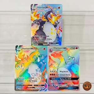 Puede incluir: Tres cartas coleccionables con Charizard. La carta superior es un Charizard VMAX negro con 330 HP. Abajo a la izquierda, un Charizard VMAX arcoíris con 330 HP. Abajo a la derecha, un Charizard GX arcoíris con 250 HP y sus ataques visibles.