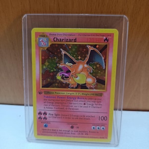 Könnte beinhalten: Eine Charizard Pokémon-Karte in einer Schutzhülle. Die Karte zeigt einen feurigen orangefarbenen Drachen mit Flügeln, einen gelben Rahmen und Text wie "Charizard" und "120 HP". Die Karte ist eine Stufe 2 Entwicklung.