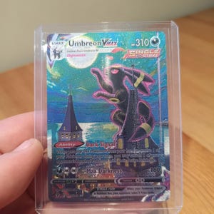 Può includere: Una carta collezionabile olografica Umbreon VMAX in una custodia protettiva in plastica trasparente. La carta raffigura un grande personaggio Umbreon nero appollaiato su un alto edificio sotto una luna piena, con toni di cielo blu e viola.