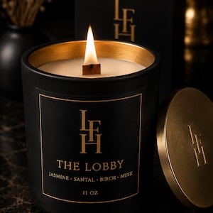 K&ouml;nnte beinhalten: Schwarze Kerze mit goldenem Rand und Deckel, beschriftet mit "THE LOBBY" und Duftnoten. Die Kerze hat einen Holzdocht und befindet sich in einer schwarzen Box. Die Kerze ist 312 Gramm.