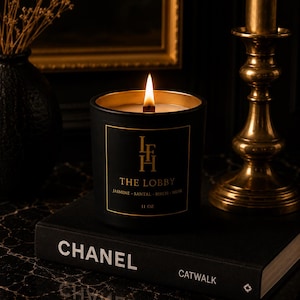 K&ouml;nnte beinhalten: Eine brennende schwarze Kerze mit goldenen Verzierungen, beschriftet mit "THE LOBBY", steht auf einem schwarzen Buch mit dem Aufdruck "CHANEL". Die Kerze wiegt 312 Gramm. Rechts befindet sich ein goldener Kerzenhalter. Links steht eine schwarze Vase mit Trockenblumen.