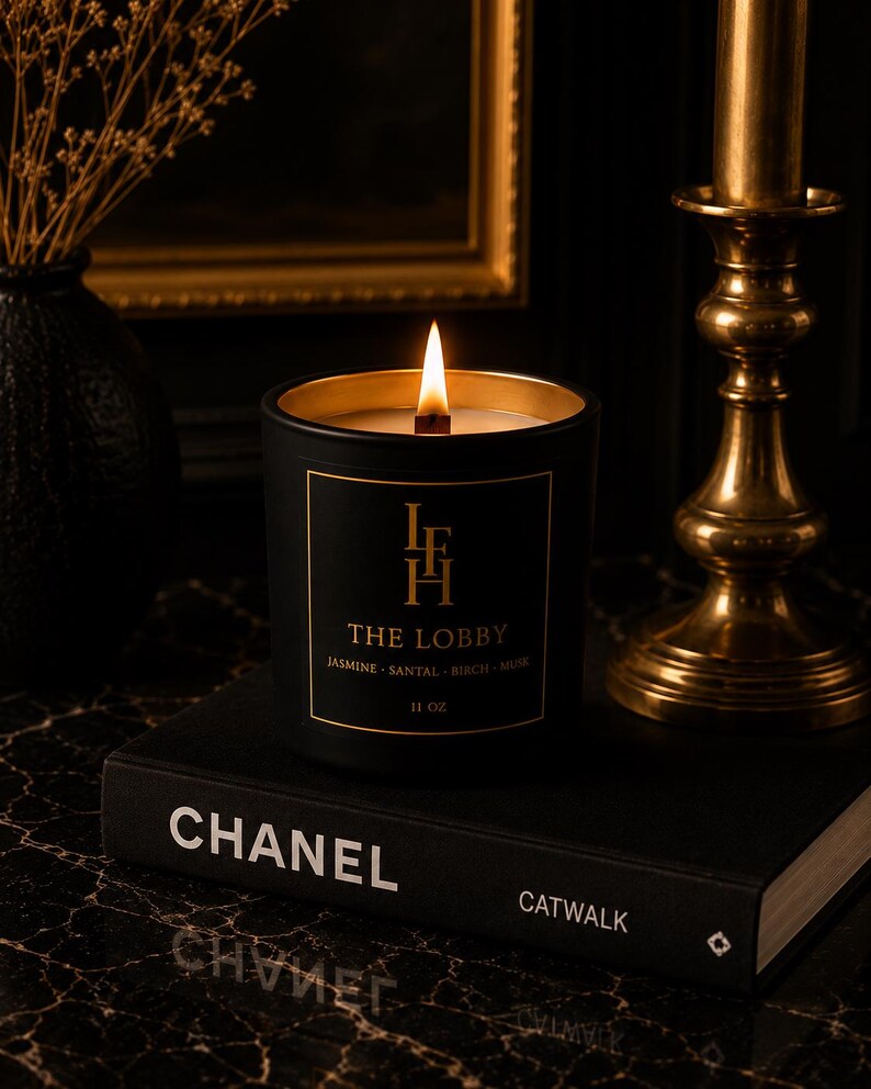 K&ouml;nnte beinhalten: Eine brennende schwarze Kerze mit goldenen Verzierungen, beschriftet mit "THE LOBBY", steht auf einem schwarzen Buch mit dem Aufdruck "CHANEL". Die Kerze wiegt 312 Gramm. Rechts befindet sich ein goldener Kerzenhalter. Links steht eine schwarze Vase mit Trockenblumen.
