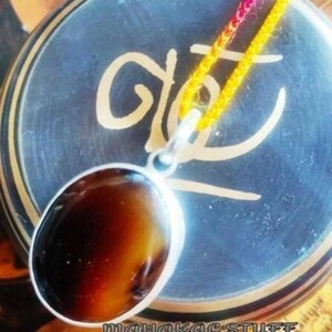 May include: An oval pendant with a brown and amber stone set in a silver-colored metal. The pendant hangs from a yellow and pink cord. The background is a dark blue surface with a gold design and the text "MAHAKAL STUFF".