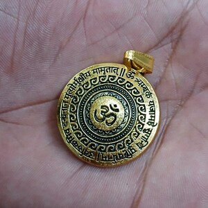 May include: A circular gold-toned pendant featuring a central Om symbol in relief. The design includes a black recessed background with intricate circular patterns and Sanskrit text inscribed along the outer edge, topped with a gold bail.