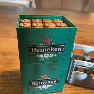 Könnte beinhalten: Ein grüner Kunststoffbehälter in Form einer Bierkiste mit dem Heineken-Logo und einem roten Stern auf der Vorderseite. Der Behälter ist mit mehreren orangefarbenen und silbernen zylindrischen Batterien gefüllt, die in Reihen angeordnet sind.