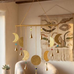 Op de afbeelding: Een goudkleurige metalen wandhanger met een horizontale stang die aan een ketting hangt. Aan de stang hangen diverse getextureerde halve maantjes en cirkelvormige elementen, afgewisseld met kleine kegelvormige metalen belletjes en kleurrijke glazen kralen.