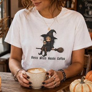 Könnte beinhalten: Ein weißes Kurzarm-T-Shirt mit einer Grafik einer Hexe mit zerzaustem Haar und spitzem Hut, die auf einem Besenstiel sitzt und eine Kaffeetasse hält. Unter der Grafik steht der Text Basic Witch Needs Coffee in schwarzer Schrift.