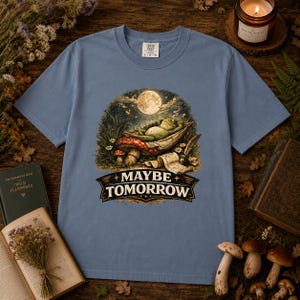 May include: A dusty blue t-shirt featuring a graphic of a frog resting in a hammock made of a mushroom under a full moon. Below the illustration, a banner displays the text "MAYBE TOMORROW" in a bold, vintage-style font with a quill and inkwell.