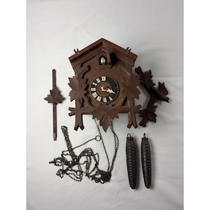 Puede incluir: Un reloj de cuco de madera marrón oscuro con un marco en forma de casa con detalles de hojas talladas, una esfera negra circular con números romanos dorados, un pequeño pájaro en una abertura superior, dos pesas en forma de piña y una cadena de metal.