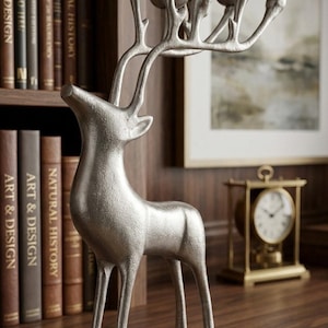 May include: Silver-colored deer-shaped candle holder with multiple candle cups. The deer figurine is standing on a wooden surface, with a bookshelf and a framed picture in the background. The candle holder is a decorative item.