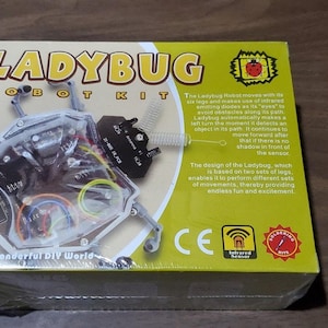 Op de afbeelding: Een verpakte Ladybug Robot Kit. De gele en witte doos toont de tekst "LADYBUG ROBOT KIT". De robot is zichtbaar door een transparant venster. De doos bevat ook tekst en een diagram van de robot.