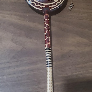 May include: A handcrafted percussion instrument with a round, dark brown head featuring an elephant design. The handle is painted with giraffe and zebra patterns. The instrument is designed for musical performances.