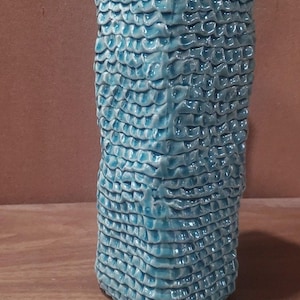 May include: A tall, textured ceramic vase in a vibrant turquoise hue. The surface is covered in a repeating, scale-like pattern, giving it a unique, tactile appearance. The vase is a decorative item.