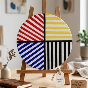 May include: A round, hand-painted artwork titled "Geometric Four" is displayed on a wooden easel. The artwork is divided into four quadrants with red and white diagonal stripes, yellow and white horizontal stripes, blue and white diagonal stripes, and black and white vertical stripes.
