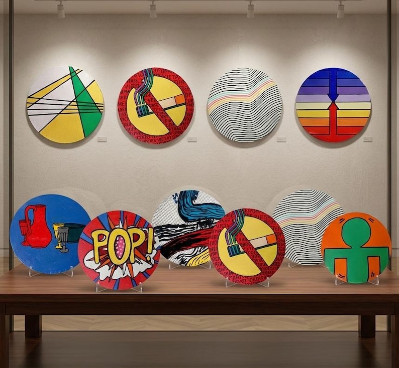May include: A collection of colorful, round decorative art pieces displayed on a wooden table. The art features various designs, including geometric shapes, pop art, and a no-smoking symbol. The pieces are mounted on clear stands.