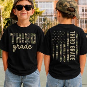 May include: A black t-shirt featuring a camouflage pattern on the front with the text THIRD grade. The back of the shirt displays a vertical American flag design in camouflage colors with the text THIRD GRADE printed in a bold, block font.