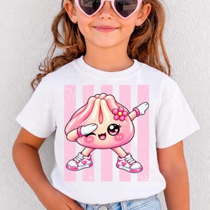 Könnte beinhalten: Ein weißes T-Shirt mit kurzen Ärmeln und einer Grafik eines rosa Cartoon-Knödels, der eine Dab-Pose einnimmt. Der Knödel hat große Augen, eine kleine Blumenverzierung und trägt weiß-rosa Turnschuhe vor einem Hintergrund mit rosa Streifen.