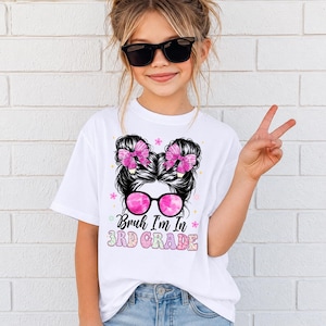 May include: A white t-shirt featuring a graphic of a person with a messy bun hairstyle, pink bows, and pink-tinted sunglasses. The text below the graphic reads "Bruh I'm In 3RD GRADE" in colorful, patterned lettering surrounded by small flower icons.