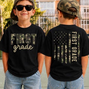 May include: A black short-sleeve t-shirt featuring a camouflage print design. The front displays the text "FIRST grade" in a blocky, camo-filled font. The back features a vertical American flag graphic in camouflage with the text "FIRST GRADE".