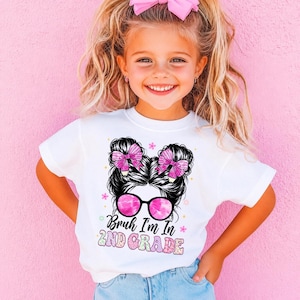 May include: A white short-sleeved t-shirt featuring a graphic of a person with a messy bun hairstyle, pink bows, and pink-tinted sunglasses. The text reads Bruh I'm In 2ND GRADE in a mix of black script and colorful patterned block lettering.