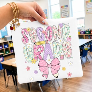 Può includere: Una pochette bianca con la scritta SECOND GRADE in motivi floreali e a pois colorati. Il design include una matita gialla, un fiocco rosa e icone di fiori. È visibile un braccialetto dorato con ciondoli a forma di mela, matita e libro.
